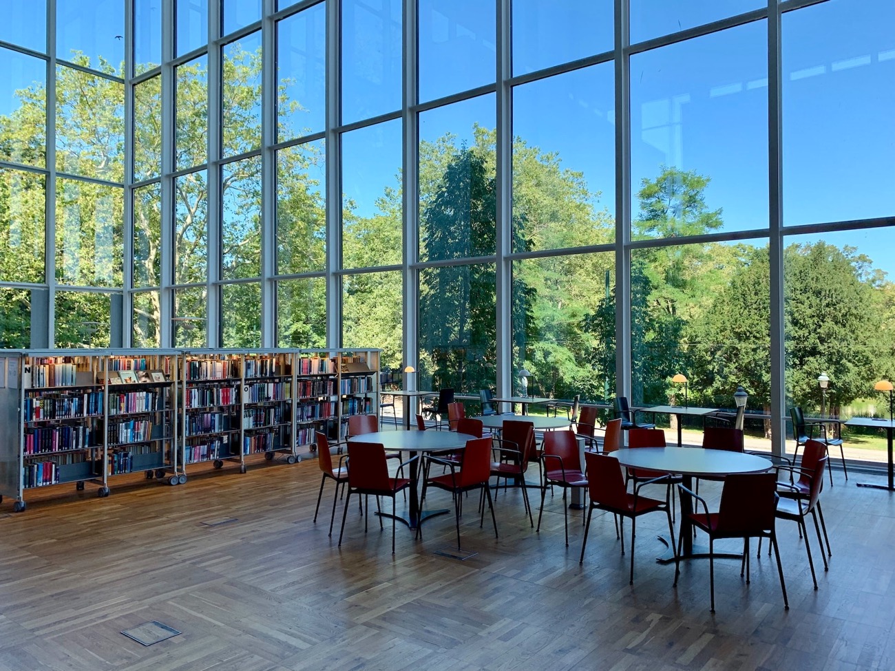 Heller Lesesaal mit Holzboden, Bücherregalen, roten Tischen und Stühlen und großen Fenstern mit Blick auf Bäume.