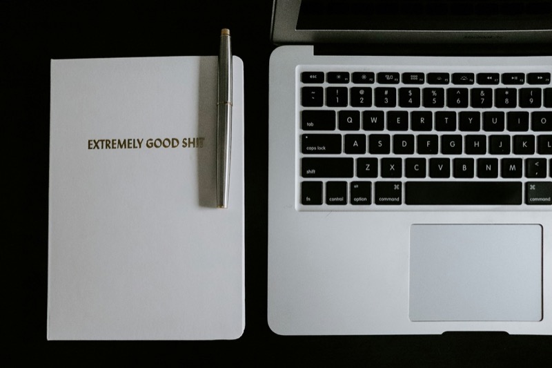 Notizbuch mit "EXTREMELY GOOD SHIT" Text, Stift und Laptop-Tastatur auf schwarzem Hintergrund.