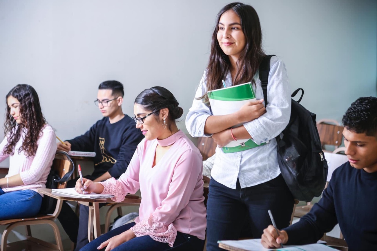 Studentin mit Rucksack und Notizbüchern steht lächelnd in einem Klassenzimmer voller sitzender Mitschüler.
